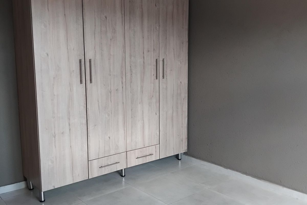 A clean, empty bedroom featuring light wood-finish built-in wardrobes, neutral grey walls, and modern tiled flooring.