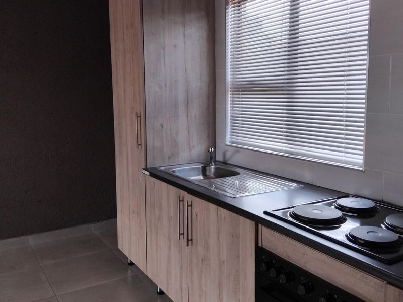 A modern, compact kitchen featuring light wood-finish cabinetry, a built-in oven and hob, stainless steel sink, and neutral tiled flooring.