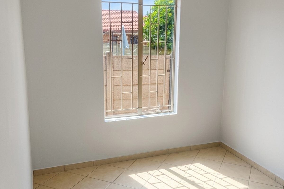 A bright, empty bedroom featuring neutral wall paint, tiled flooring, and a window fitted with security bars.