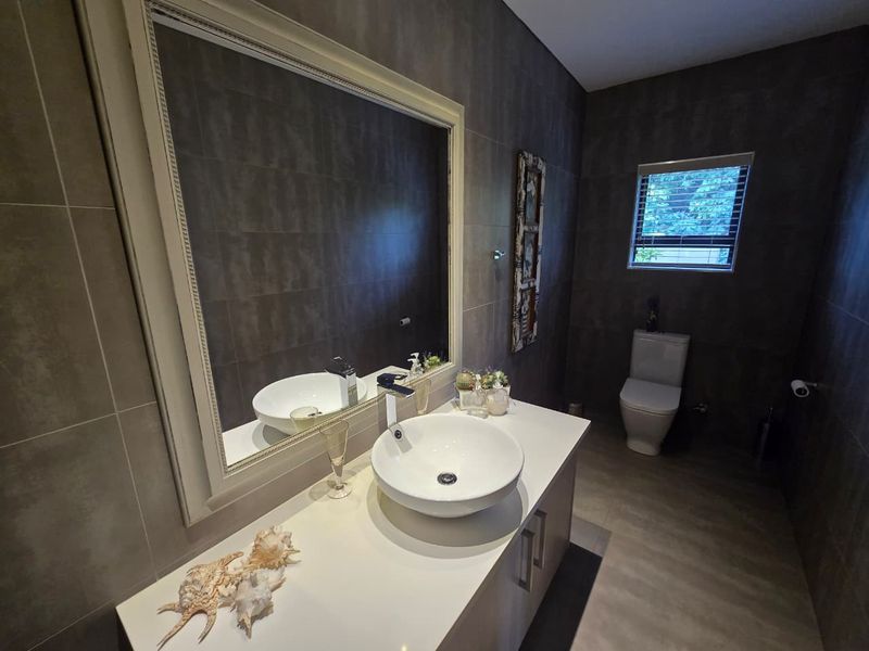 Modern bathroom featuring dark wall tiles, a sleek vanity with a countertop basin, a large framed mirror, and a private toilet area.