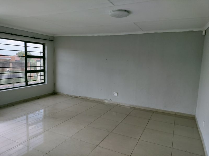 A bright, empty living room featuring neutral tiled flooring, large windows with security bars, and a simple ceiling-mounted light fixture.