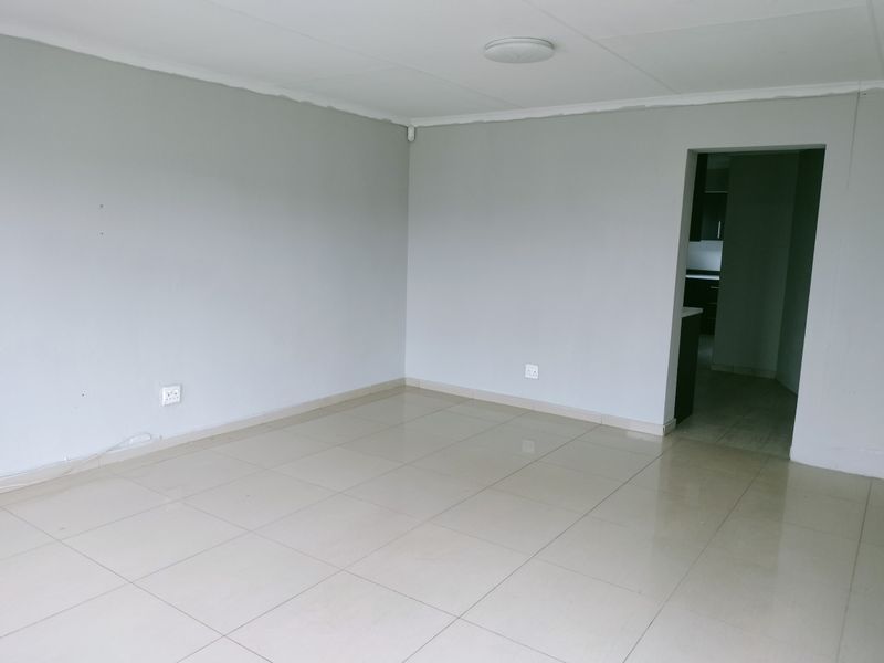 A bright, empty living area featuring neutral grey walls and light-colored floor tiles, with a direct view into the adjacent kitchen space.