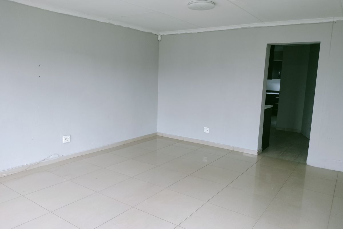 A bright, empty living area featuring neutral grey walls and light-colored floor tiles, with a direct view into the adjacent kitchen space.