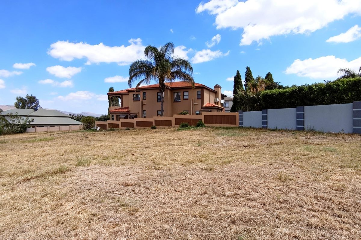 A view of a vacant residential plot featuring dry grass, perimeter boundary walls, and a neighboring multi-story residential property.