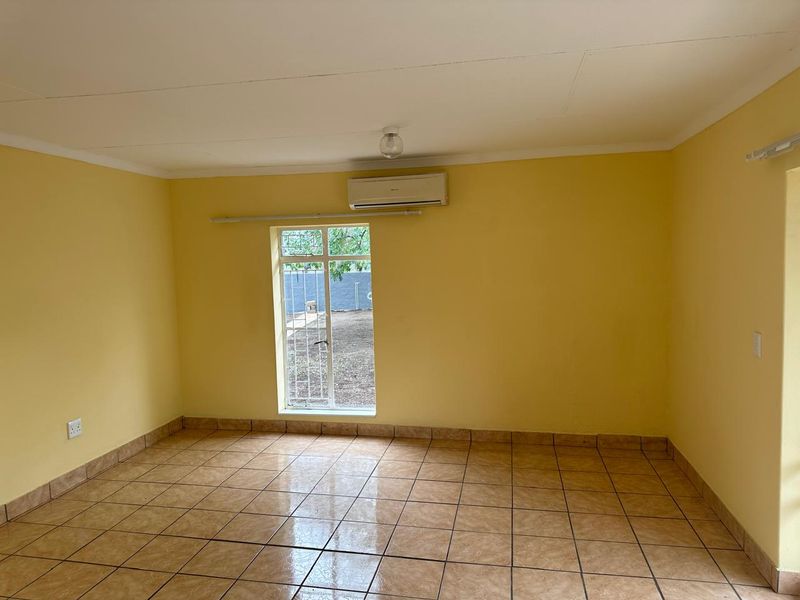 An empty living room featuring tiled flooring, a wall-mounted air conditioning unit, and a window with security bars.