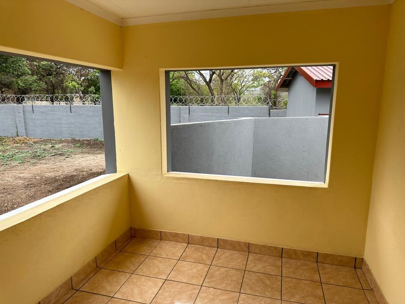 An empty, covered patio area featuring tiled flooring and large window openings looking out onto the rear garden and boundary wall.