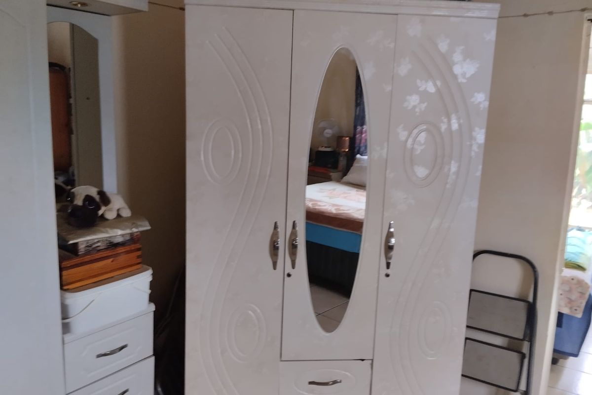 A bedroom interior featuring a large freestanding wardrobe with a central mirror and integrated storage drawers.