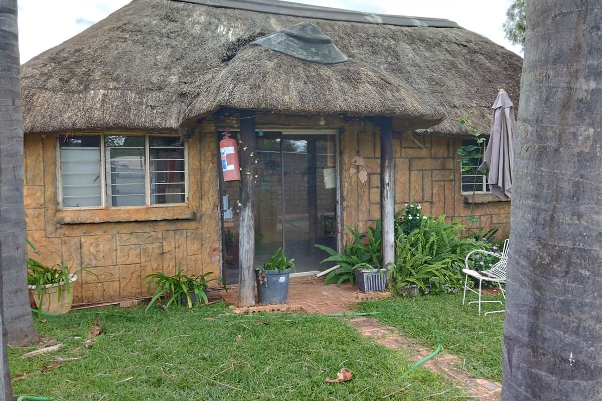 Exterior view of a rustic cottage featuring a traditional thatched roof, textured wall cladding, and sliding glass entrance doors set within a garden.