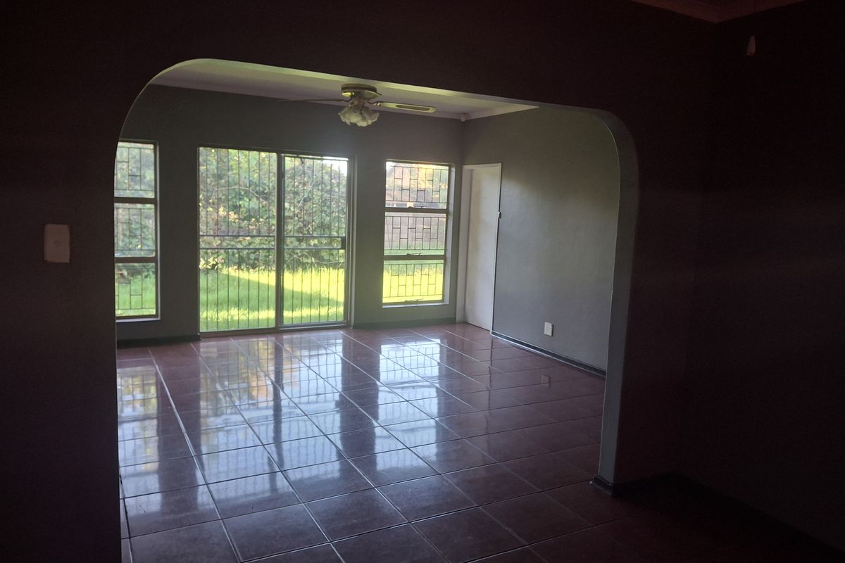 A spacious, empty living area featuring tiled flooring, an arched entryway, and large windows with security bars overlooking the garden.