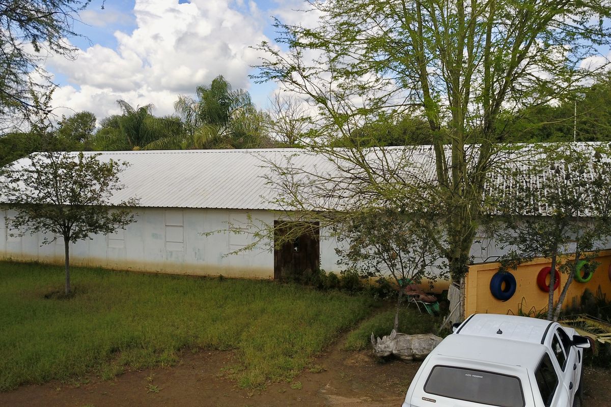 Exterior view of a long, single-story building with a corrugated metal roof, set within a rural small holding landscape.