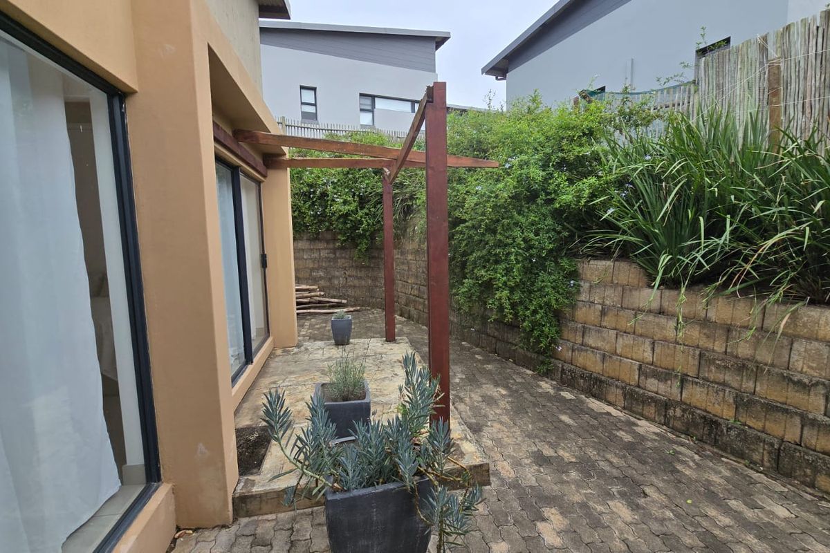 A rear patio area featuring a timber pergola structure, paved flooring, and a tiered retaining wall with established garden greenery.
