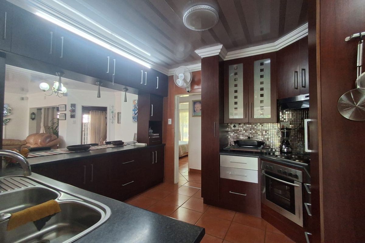 A functional kitchen featuring dark wood cabinetry, a breakfast counter, built-in oven with hob, and mosaic tile backsplash, opening into the living area.