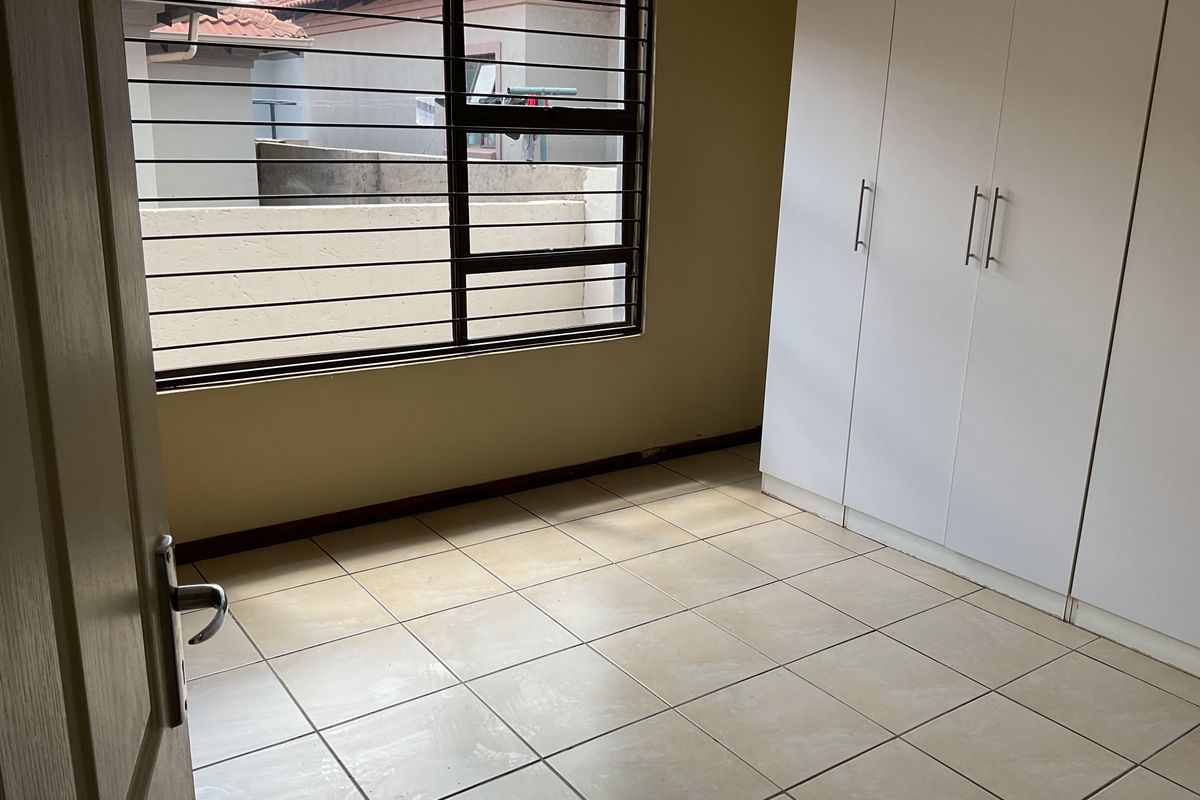 A vacant bedroom featuring neutral tiled flooring, built-in white cupboards, and a window fitted with security bars.