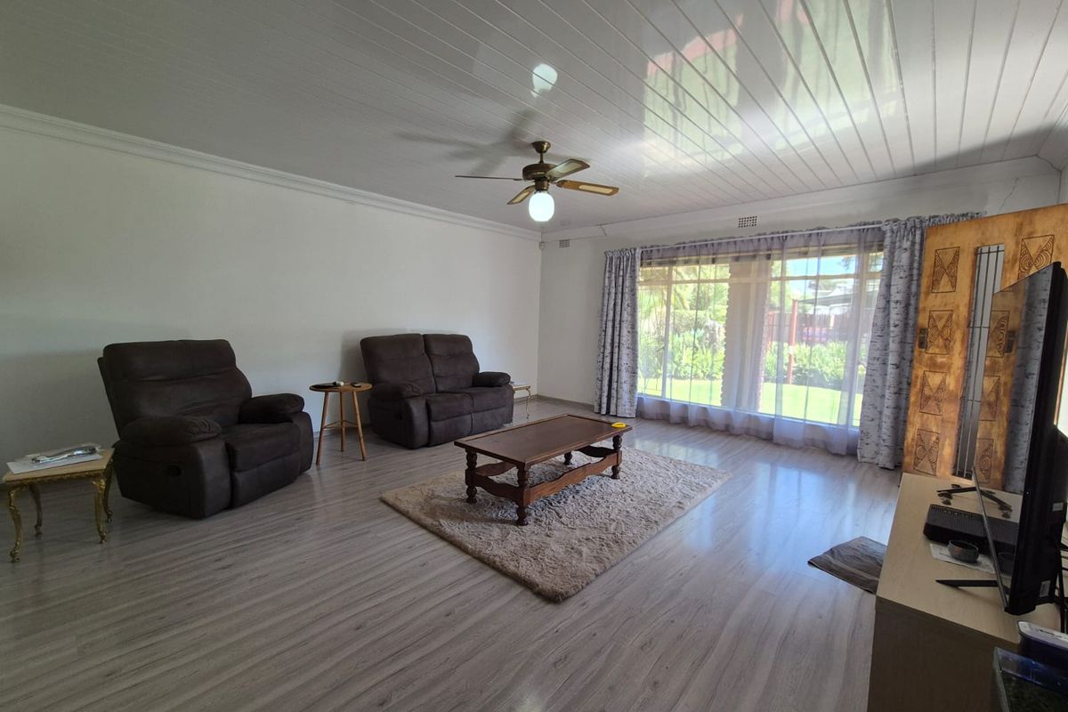 A spacious living room featuring light-toned laminate flooring, a white tongue-and-groove ceiling, and a ceiling fan, with large windows overlooking the garden.