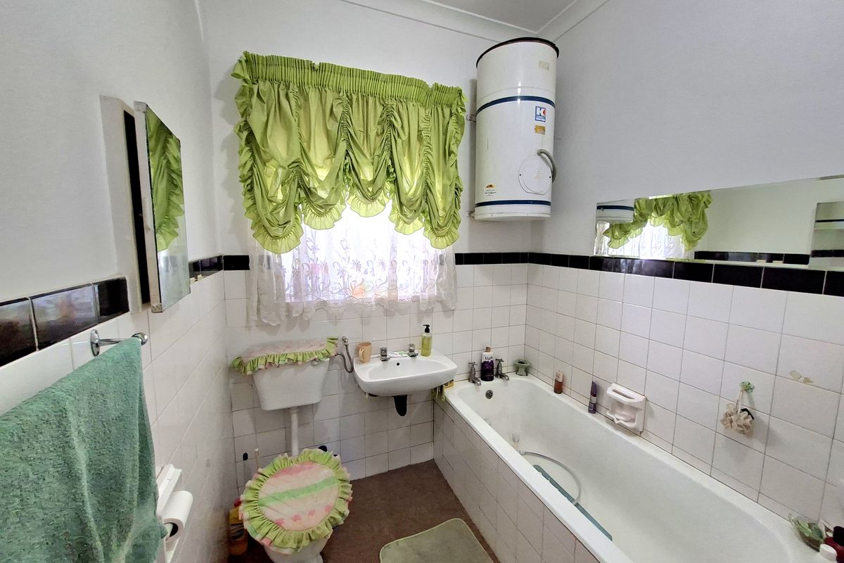 A compact bathroom featuring a standard bathtub, wall-mounted basin, and an exposed electric water heater, finished with dated white wall tiles.
