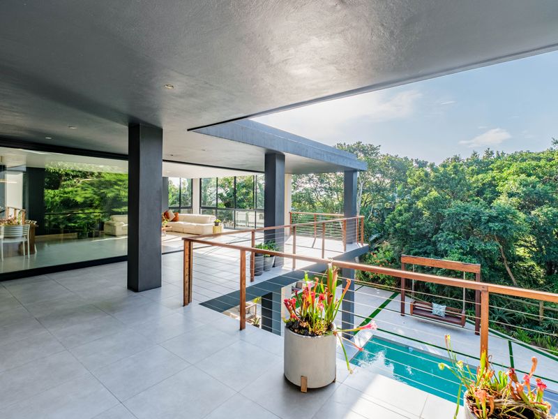 Spacious covered balcony featuring modern tiled flooring, structural columns, and a timber balustrade, overlooking a private swimming pool and lush garden surroundings.