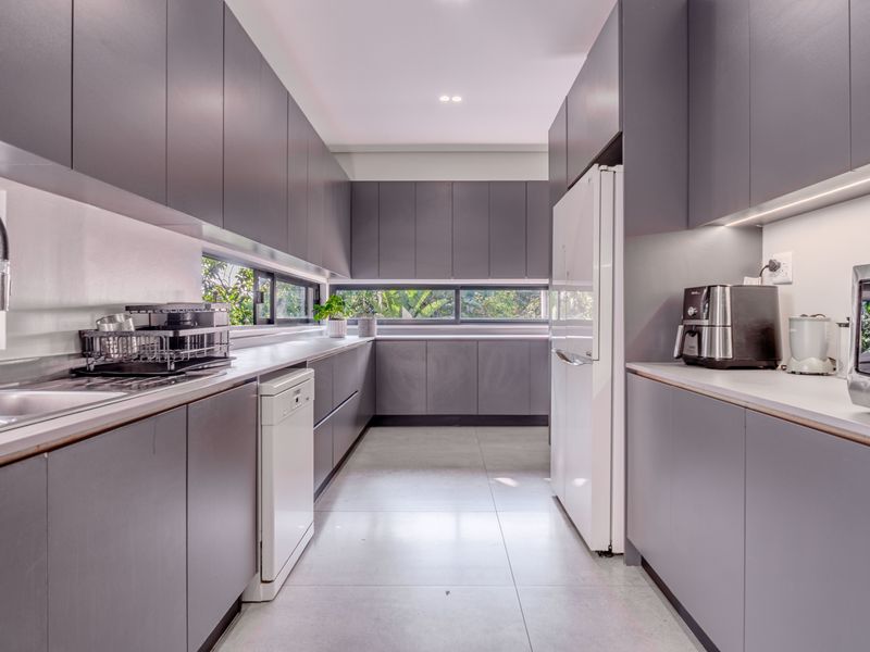 Modern galley-style kitchen featuring sleek grey cabinetry, integrated appliances, tiled flooring, and horizontal windows providing natural light and garden views.