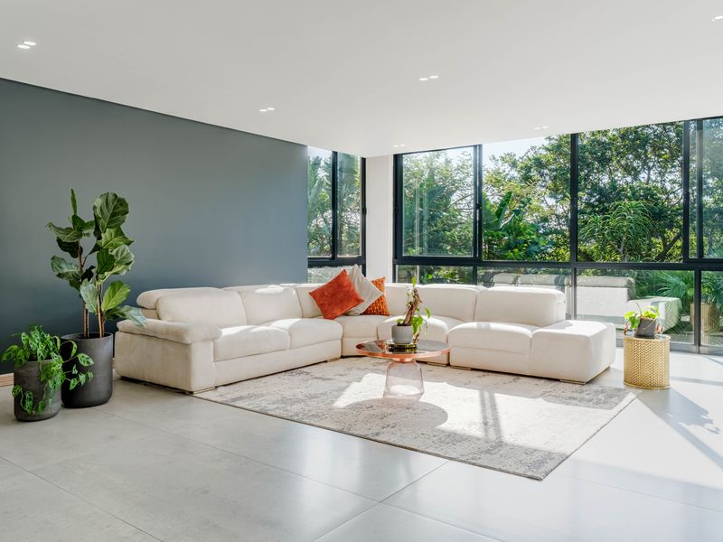 A bright and modern living room featuring large format floor tiles, a dark feature wall, and expansive aluminium windows overlooking lush greenery.