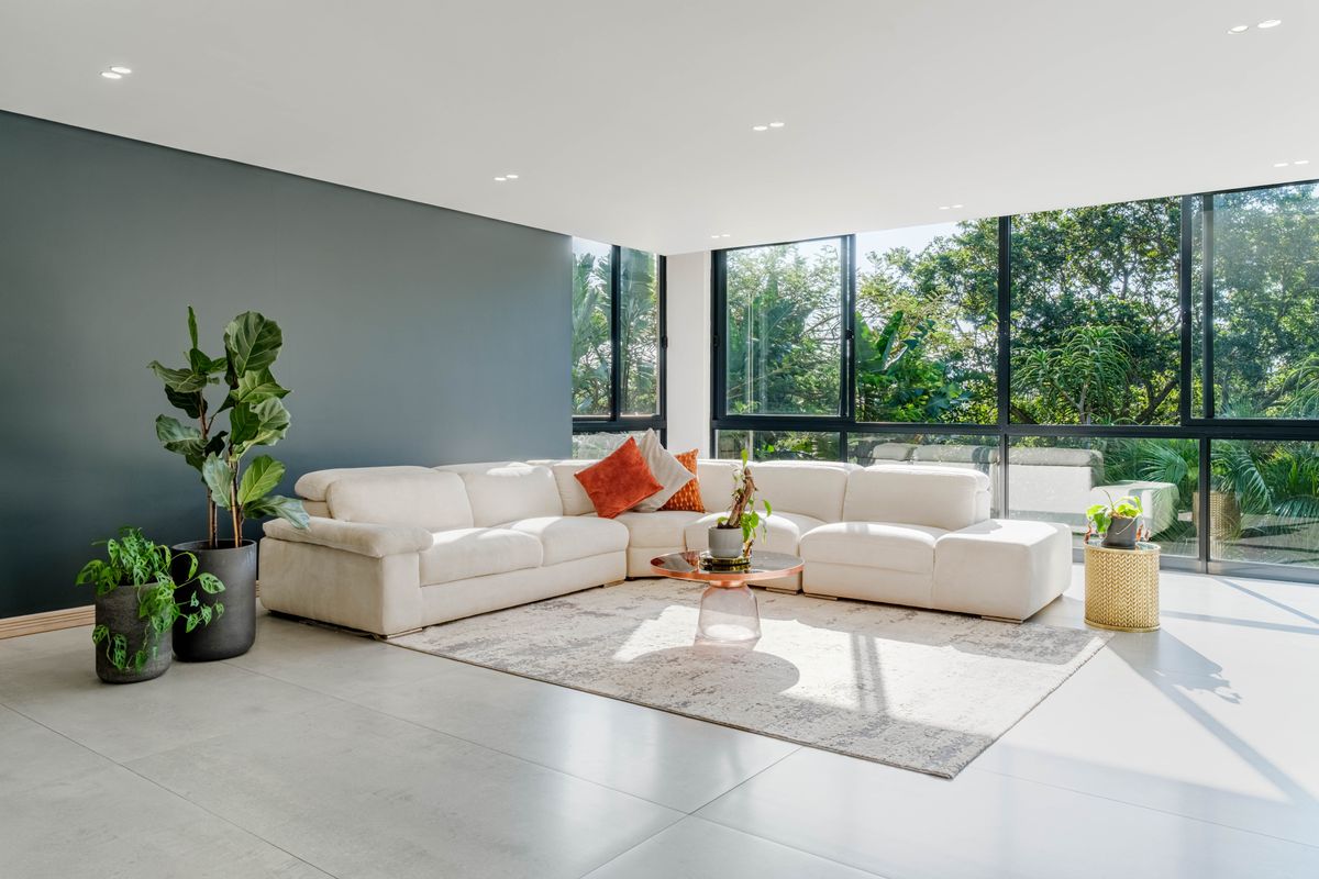 A bright and modern living room featuring large format floor tiles, a dark feature wall, and expansive aluminium windows overlooking lush greenery.