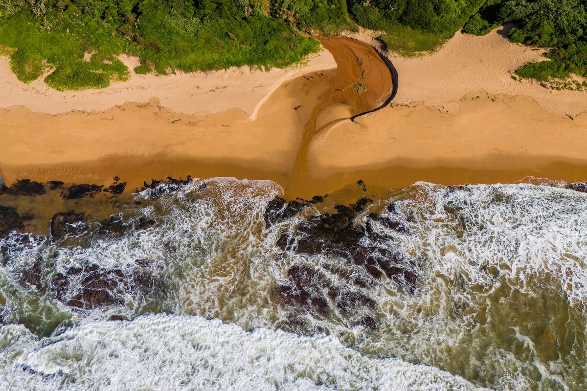 An aerial view of the coastal landscape featuring a sandy beach, rocky shoreline, and natural estuary flowing into the ocean.