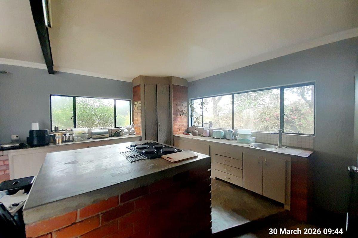 A rustic kitchen featuring a brick-based central island with a built-in hob, concrete countertops, and expansive windows overlooking the farm grounds.