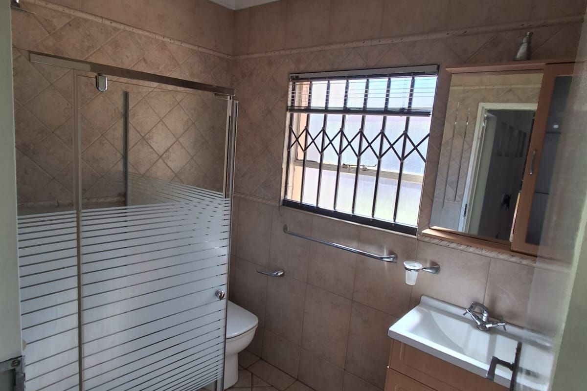 A functional bathroom featuring a glass-enclosed shower, a vanity cabinet with a basin, and a window with security bars.