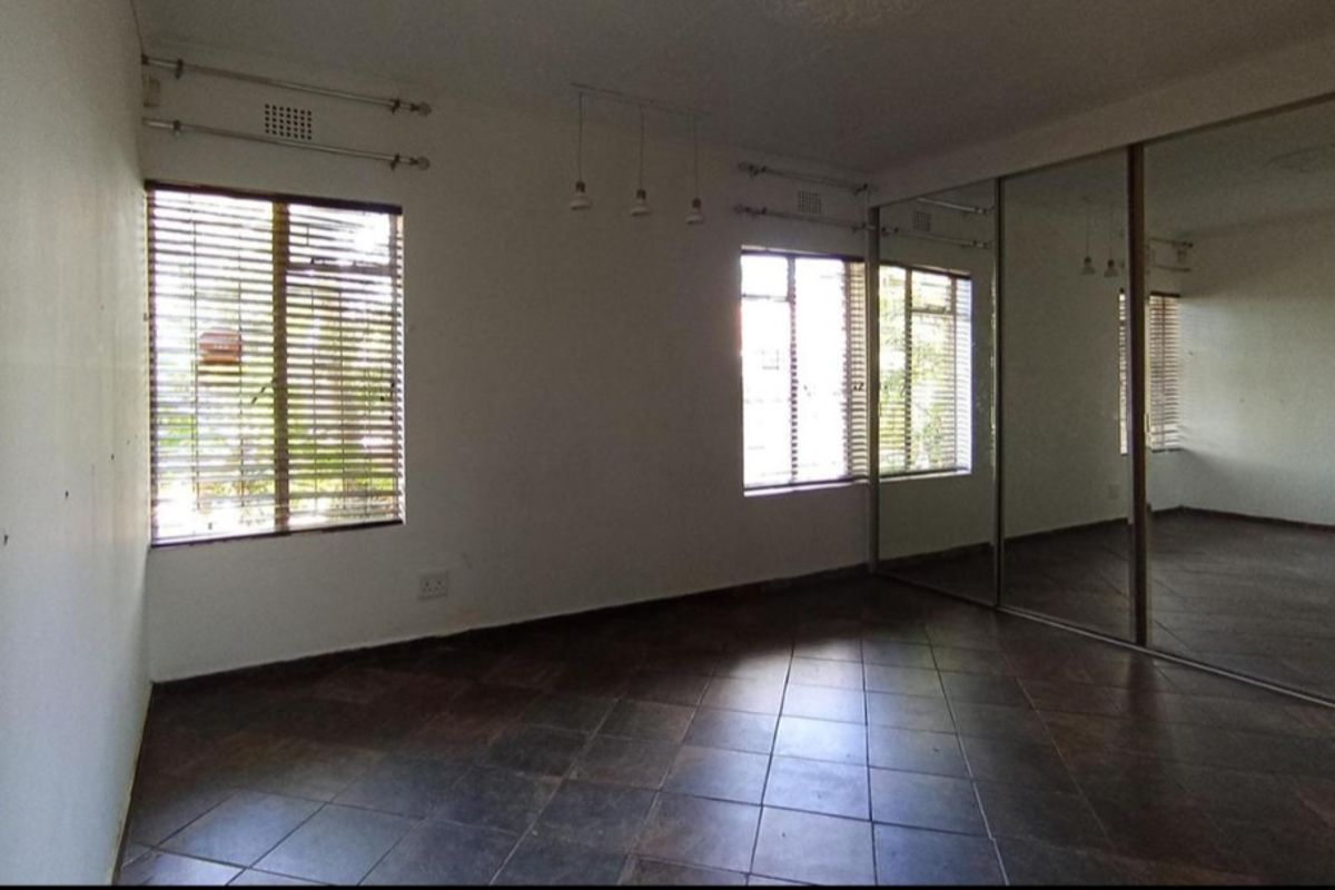 A spacious bedroom featuring dark tiled flooring, large windows with fitted blinds, and extensive built-in mirrored wardrobes.