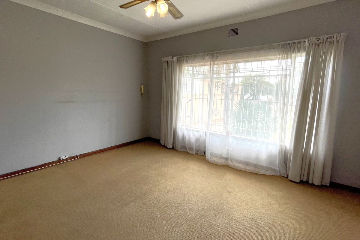 A spacious, empty bedroom featuring neutral walls, carpeted flooring, a ceiling fan, and large windows with security bars.