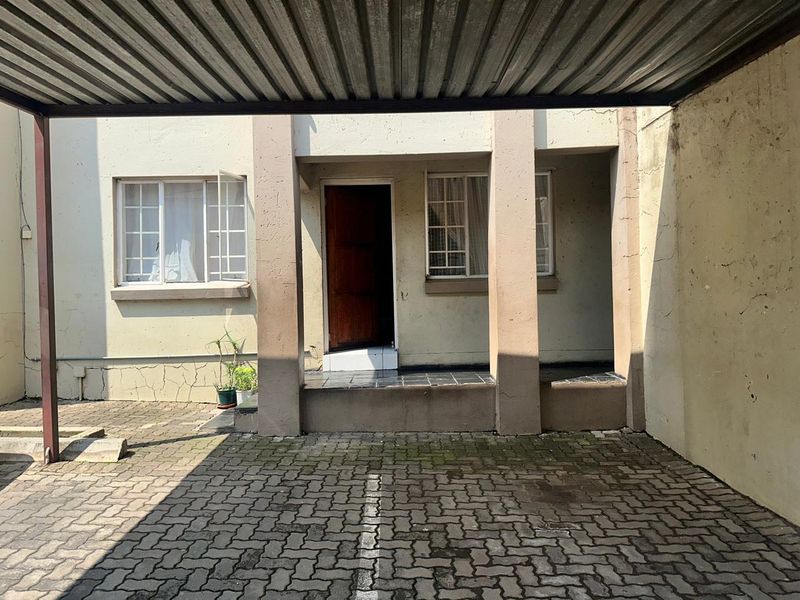 Front entrance of the flat featuring a covered carport, paved driveway, and a tiled porch area with a timber front door.