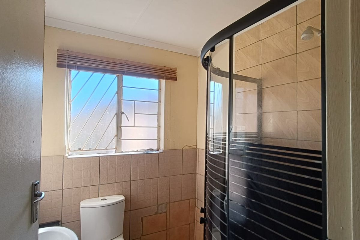 A functional bathroom featuring a corner shower enclosure, a standard toilet, and a window with security bars, requiring some cosmetic updates to wall finishes.