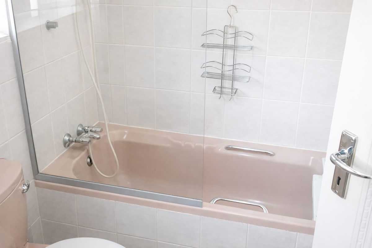 A dated bathroom featuring a peach-colored bathtub and matching toilet, complemented by white wall tiles and a glass shower screen.