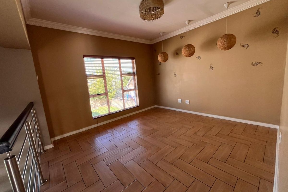 A spacious, empty living area featuring herringbone-patterned wood-look floor tiles, decorative pendant lighting, and a large window with fitted blinds.