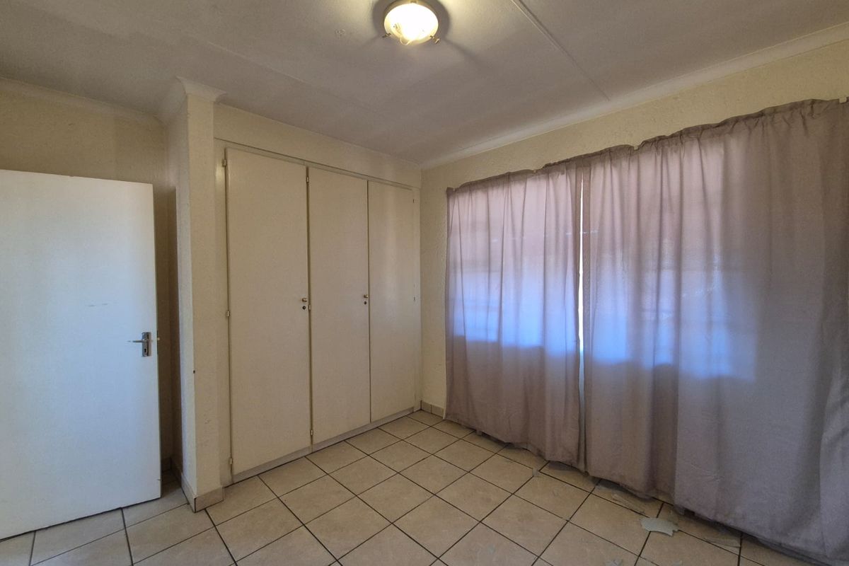 A bedroom featuring built-in cupboards, tiled flooring, and a window with curtains. The room is vacant and requires cosmetic updates.