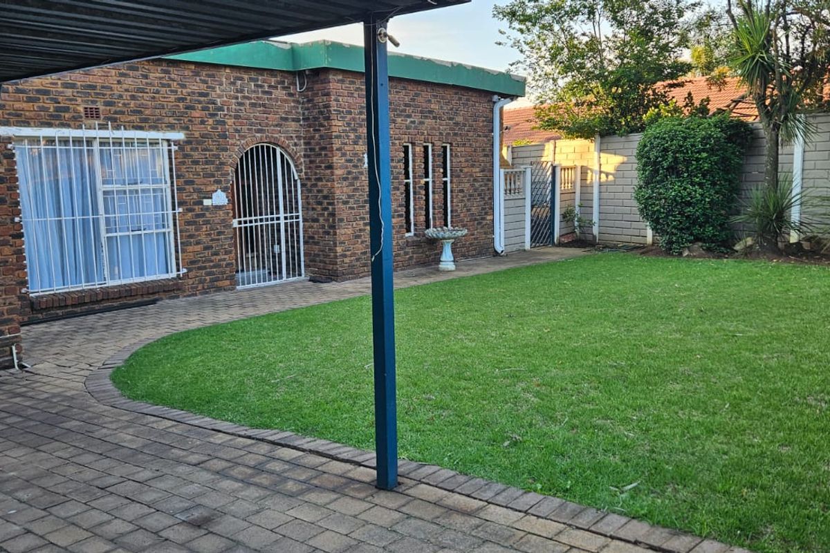 A rear view of the property featuring a brick facade, a paved patio area under a carport, and a well-maintained lawn with boundary walls.