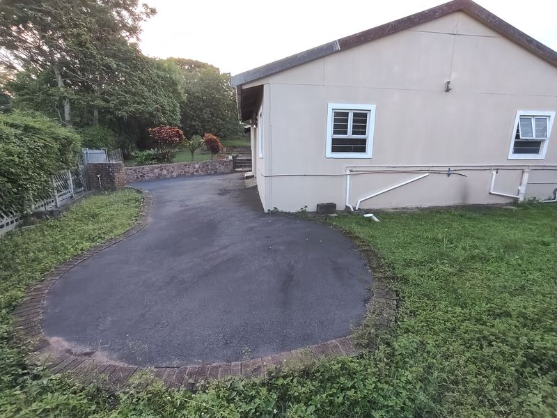 Rear exterior view showing an asphalt driveway, stone retaining wall, and landscaped garden area with visible external plumbing and window security bars.
