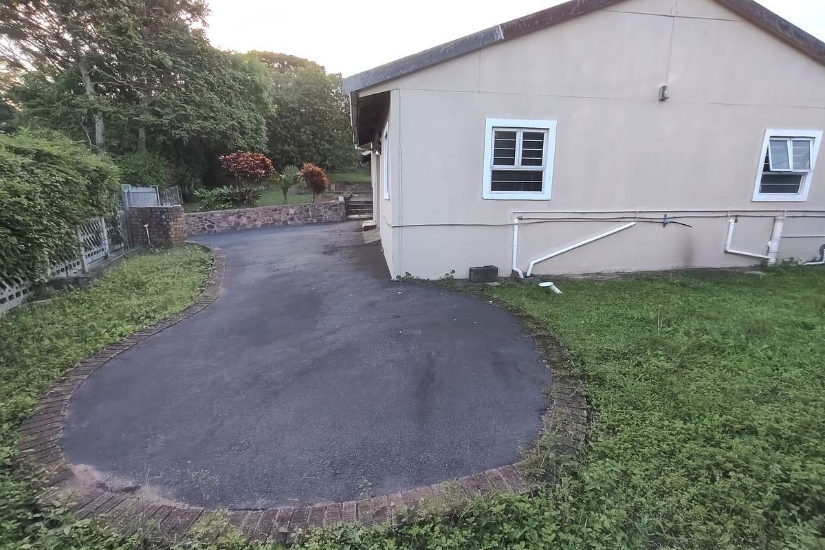 Rear exterior view showing an asphalt driveway, stone retaining wall, and landscaped garden area with visible external plumbing and window security bars.