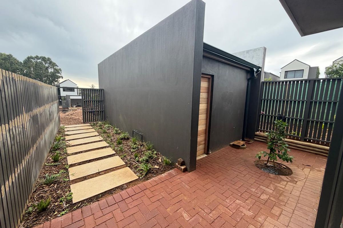 A private rear courtyard featuring brick paving, a modern dark-rendered wall, timber door, and a landscaped garden path leading to a side gate.