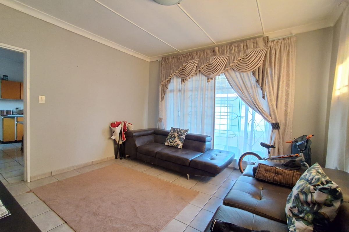 A living room featuring tiled flooring, coved ceilings, and a large window with decorative curtains, providing access to an adjacent kitchen area.
