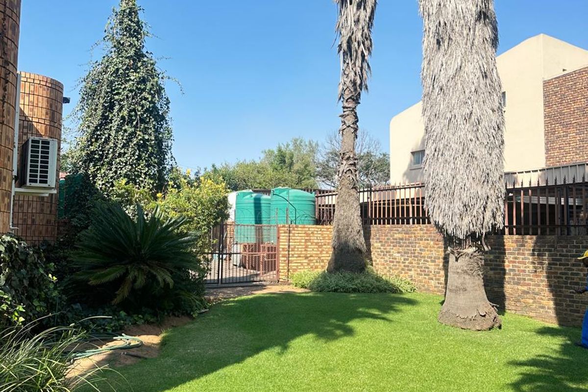 A well-maintained rear garden featuring a manicured lawn, mature palm trees, brick boundary walls, and external water storage tanks.