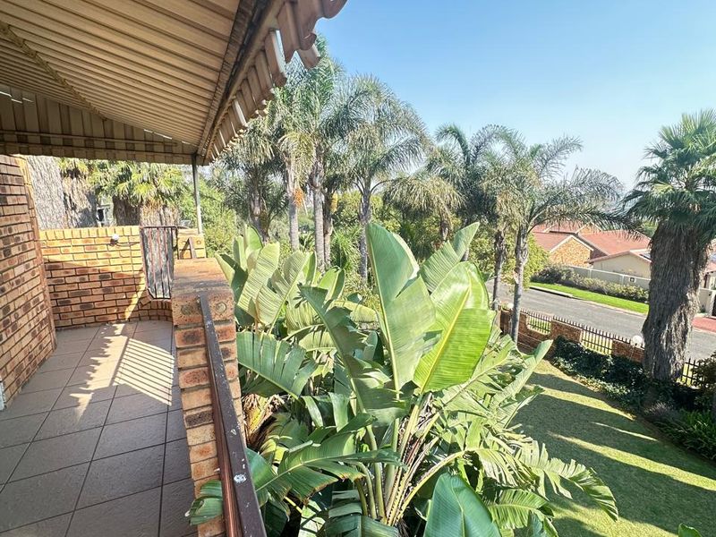 A covered balcony featuring tiled flooring and brick balustrades, overlooking a lush garden with mature palm trees and a suburban street view.