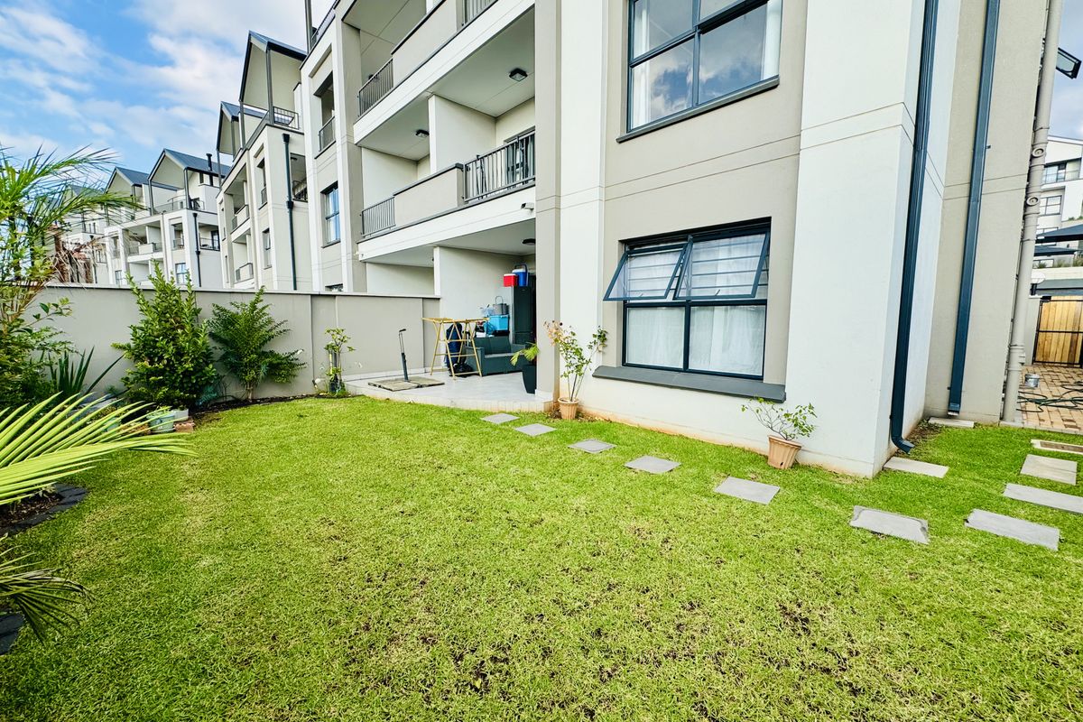A private ground-floor garden area featuring a paved patio, manicured lawn, and direct access to the modern apartment building exterior.