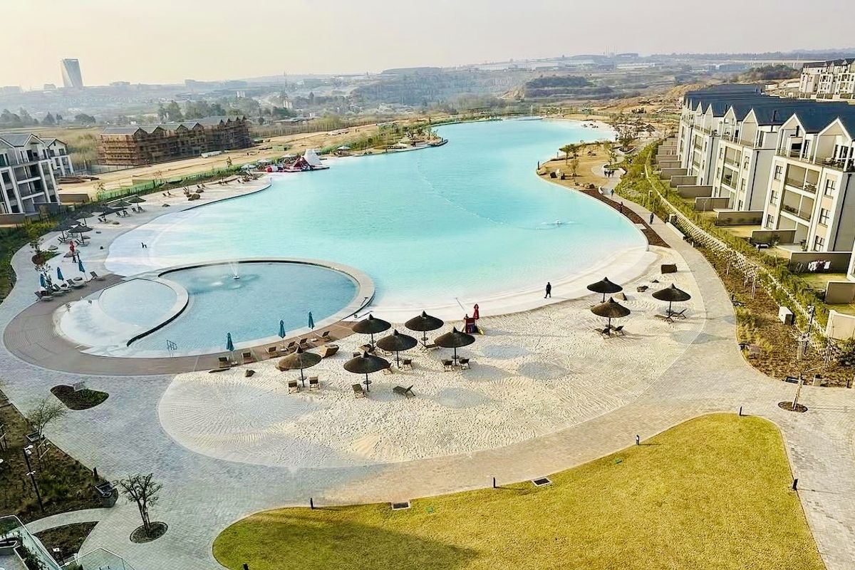 Aerial view of a large communal lagoon and beach area featuring landscaped grounds, paved walkways, and modern residential apartment blocks.