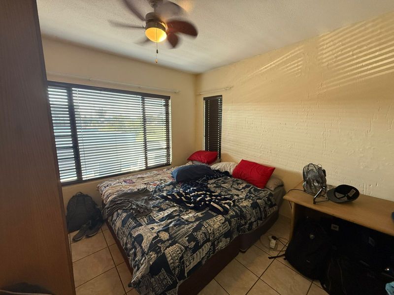 A bedroom featuring tiled flooring, a ceiling fan, and large windows with fitted blinds, offering a functional space with potential for cosmetic updates.