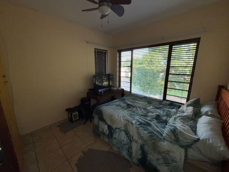 A bedroom featuring tiled flooring, a ceiling fan, and large windows with fitted blinds, providing natural light and views of the surrounding area.
