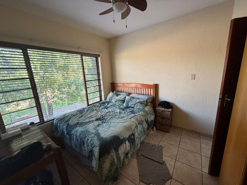 A bedroom featuring tiled flooring, a ceiling fan, and large windows with blinds, offering a functional space with natural light.