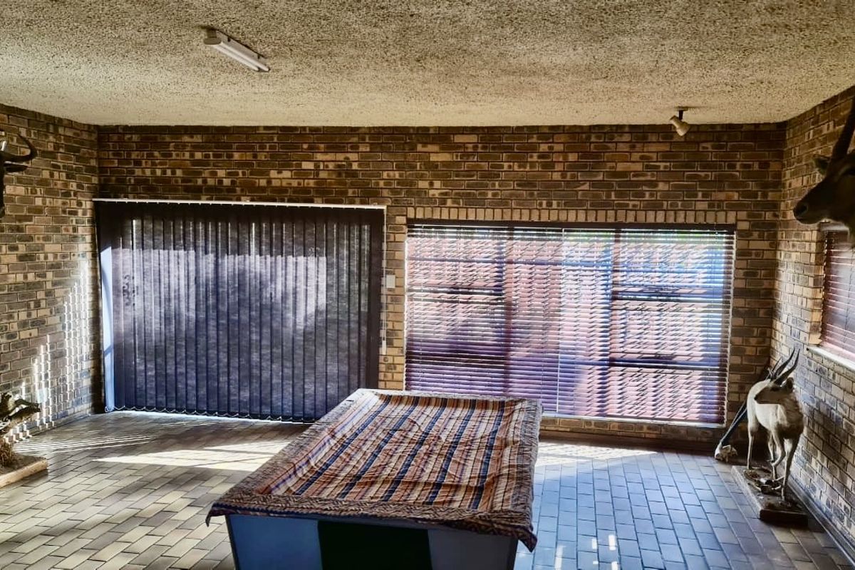 Spacious living area featuring exposed face-brick walls, tiled flooring, and large windows with fitted blinds, offering a rustic and functional entertainment space.