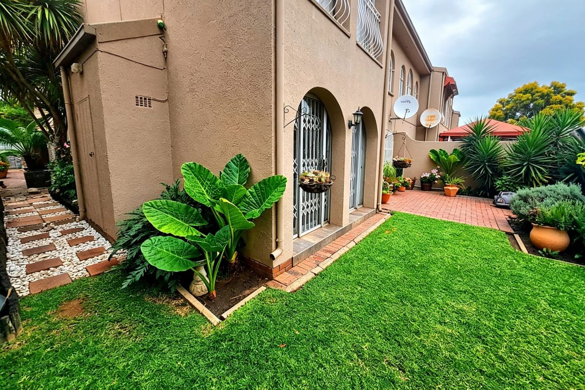 A well-maintained rear exterior view of the townhouse featuring a paved patio area, manicured lawn, and established garden beds.