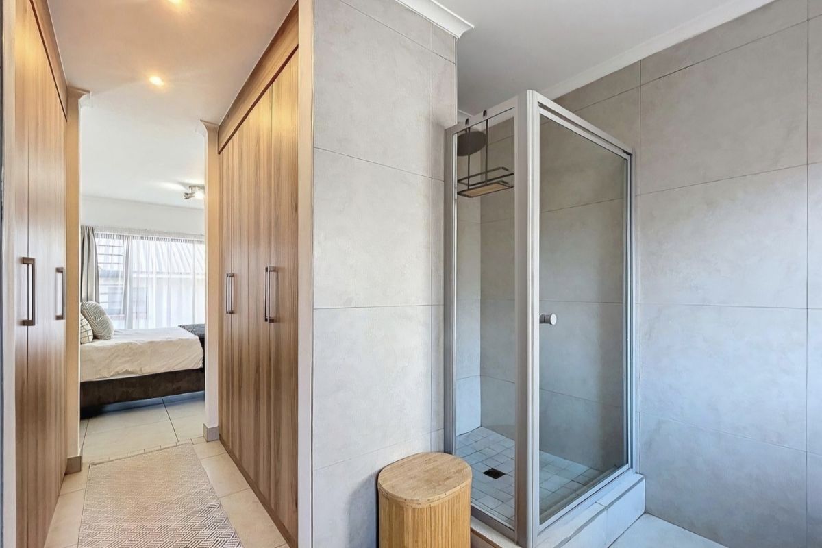 Modern bathroom featuring a glass-enclosed shower with neutral wall tiling, adjacent to built-in wooden cabinetry and a view into the bedroom area.