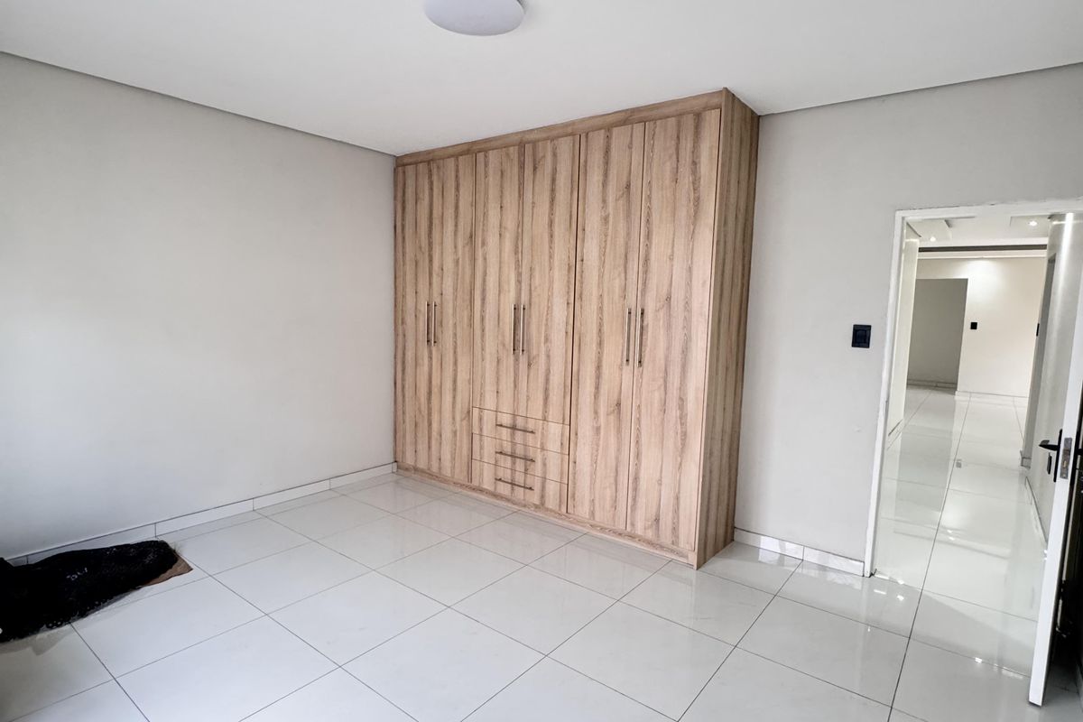 A bright, empty bedroom featuring modern light wood built-in cupboards and polished white floor tiles, leading into a hallway.