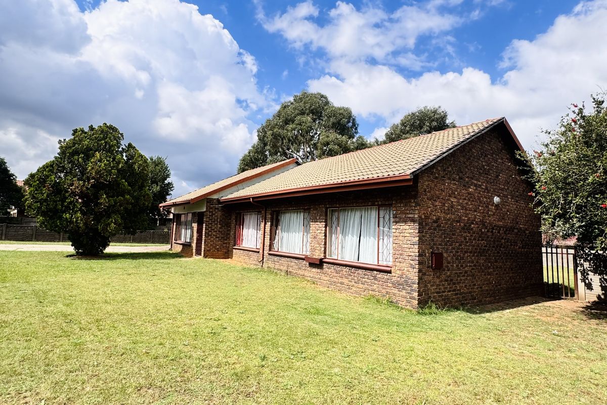 A view of the side and rear exterior of a single-story brick house set on a large, open grass lawn.